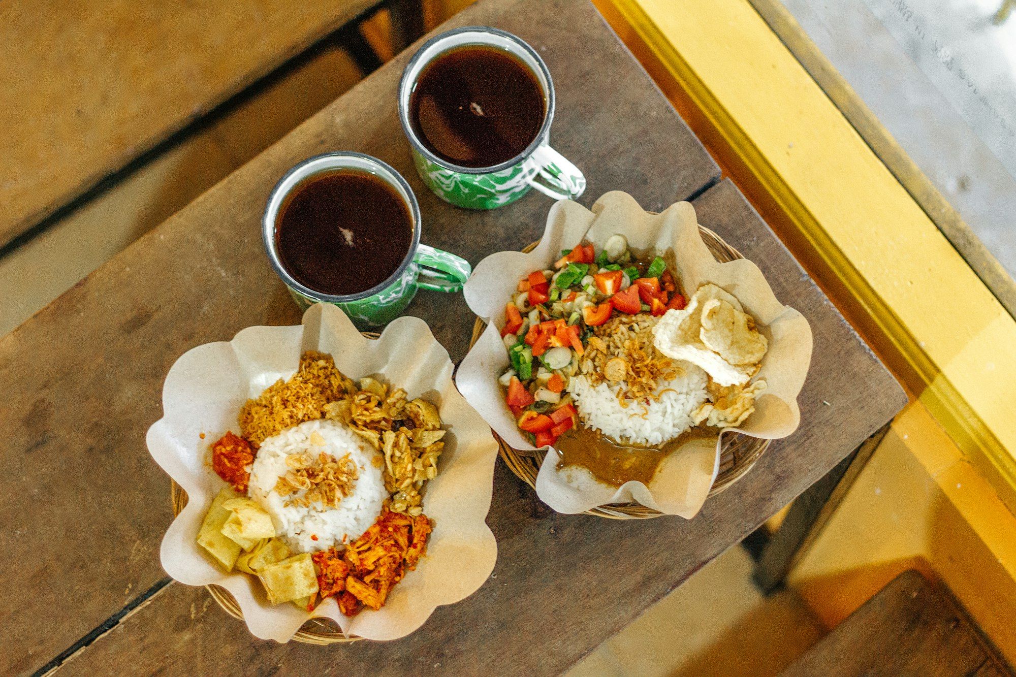 Dua porsi nasi campur di keranjang bambu beralas kertas, dengan dua gelas kopi hitam, dipotret dari atas di meja kayu