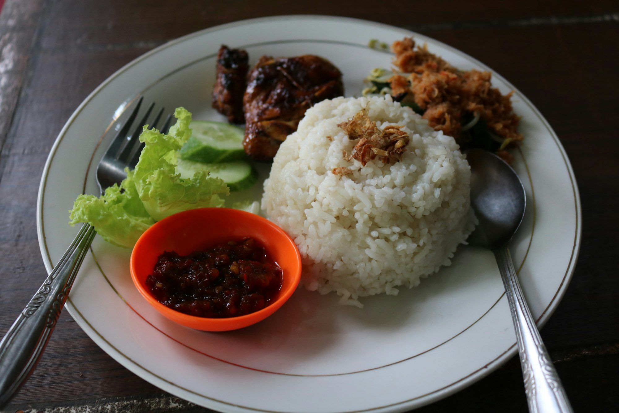 Piring nasi putih dengan ayam bakar, sambal dalam mangkuk oranye, selada, dan irisan timun, sendok garpu di kedua sisi