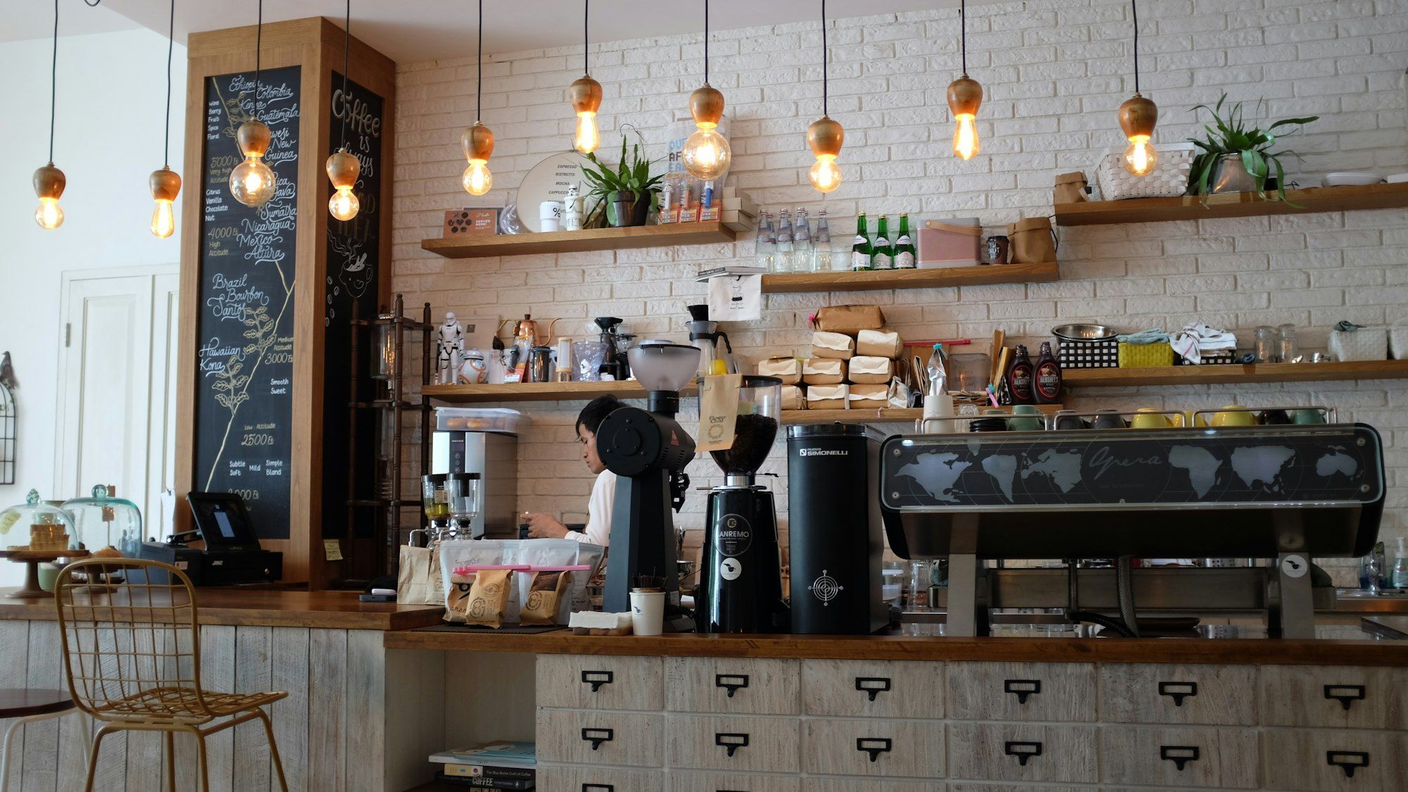 Bar depan dengan mesin espresso, papan tulis menu tinggi, lampu bohlam gantung, dan rak kayu berisi tanaman di dinding bata putih