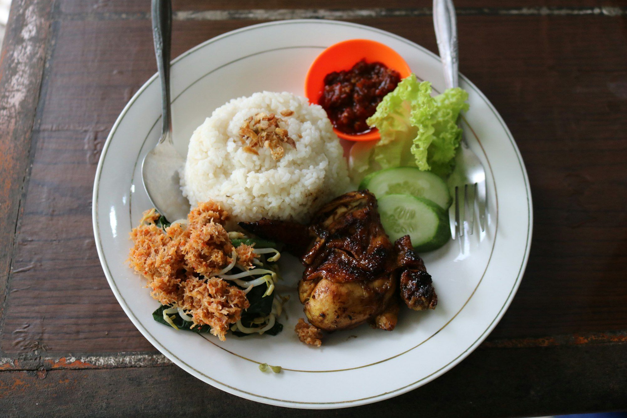 Piring nasi putih dengan ayam bakar kecap, sambal merah di mangkuk kecil oranye, selada, irisan timun, dan tumis kacang panjang
