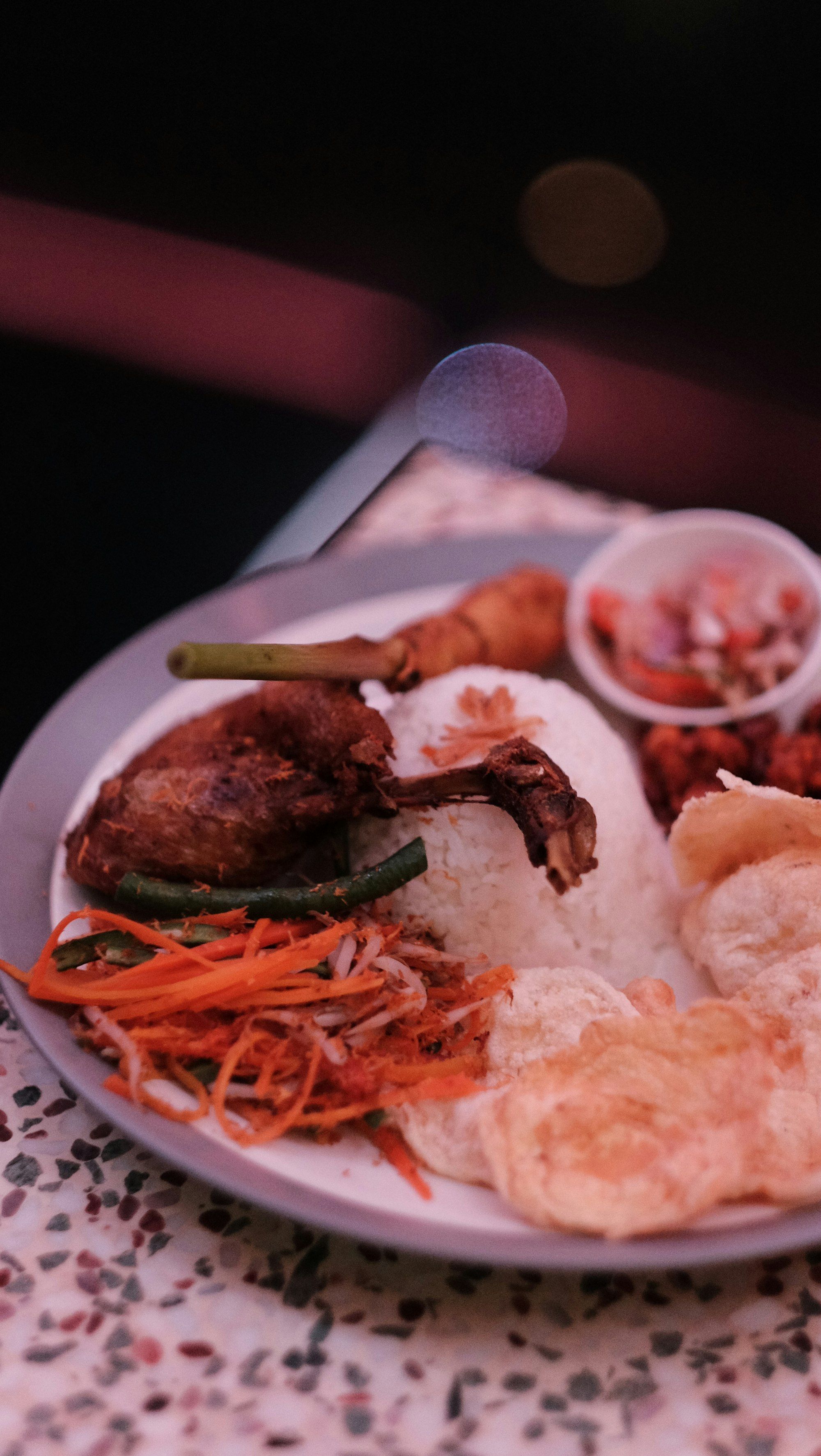 Piring nasi dengan bebek goreng utuh, kerupuk, acar wortel, sambal matah dalam mangkuk plastik, di atas meja teraso