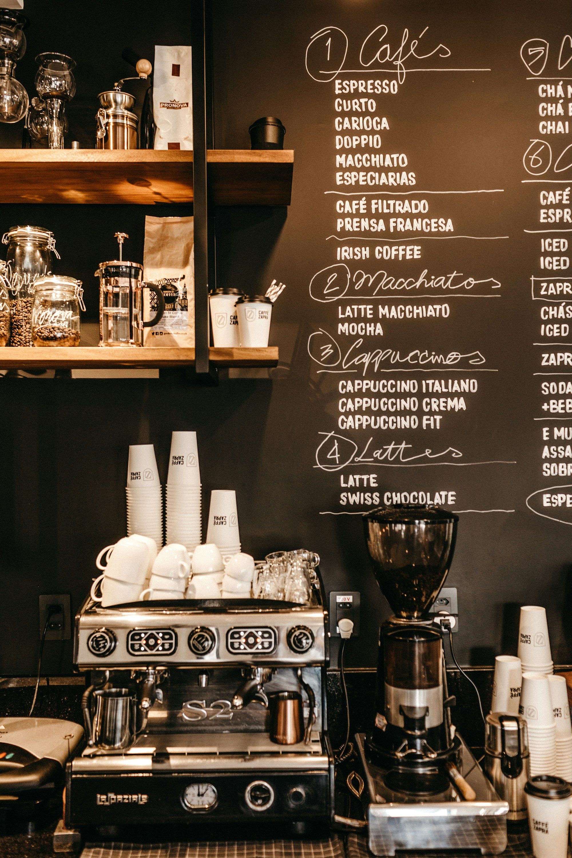 Papan tulis hitam berisi daftar menu tulisan tangan yang dinomori, di bawahnya mesin espresso dan tumpukan gelas