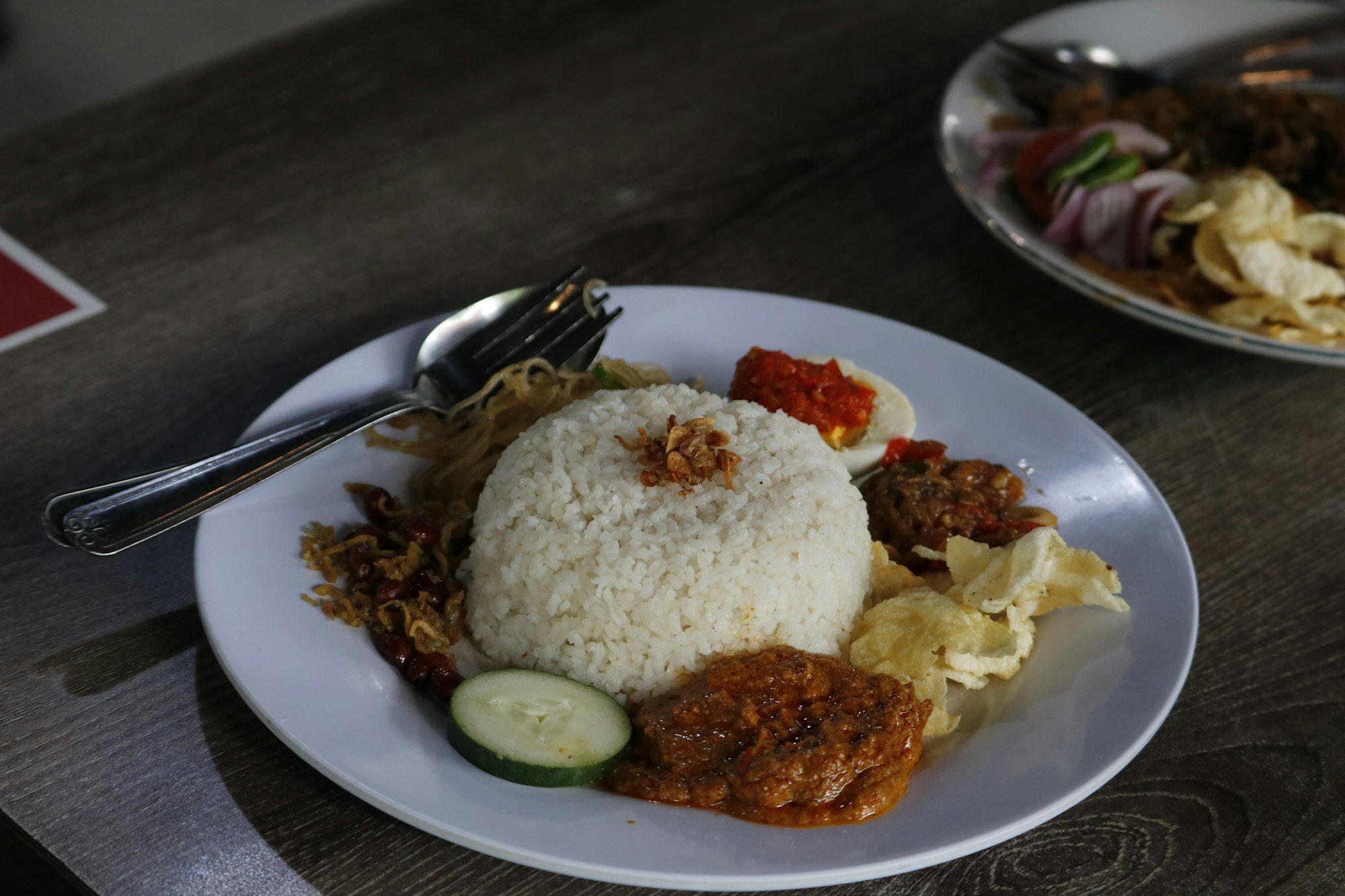 Piring nasi campur dengan sambal merah, telur, kerupuk, dan irisan timun, dengan piring kedua yang samar di belakangnya